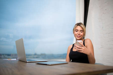 Young hipster girl reading text message on mobile phone, sitting at table with laptop computer and...