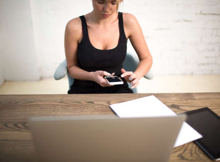 Hipster girl typing text on mobile phone, sitting at the wooden table with laptop computer, touch...