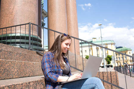 Cheerful woman student preparing for the course work using modern laptop computer. Smiling female tourist or freelancer chatting on net-book, sitting outdoors near university or architectural buildingの写真素材
