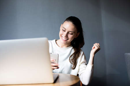 Happy woman writer received good news in text message on mobile phone during work on laptop computer. Smiling female student reading positive sms on her cell telephone, sitting with net-book in cafeの写真素材