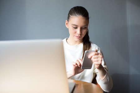 Charming hipster girl typing text on mobile phone after distance work on laptop computer. Pretty...