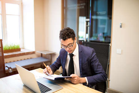 Successful businessman reading information in notepad while sitting at the table with laptop...