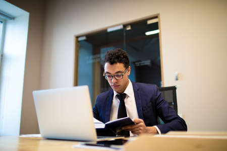 Professional Afro businessman in formal suit reading information in textbook, sitting at table with...