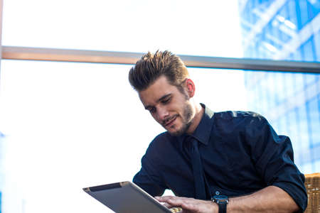 Smiling man entrepreneur online payment via digital tablet, sitting in business center near window with copy space. Handsome male employer using messenger on touch pad during work break in companyの写真素材