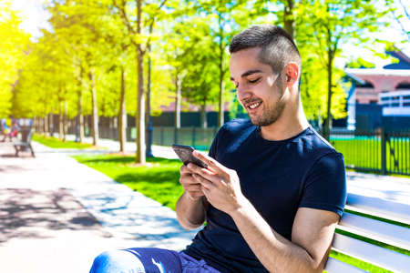 Smiling hipster guy playing games in social network via mobile phone while relaxing in city park...
