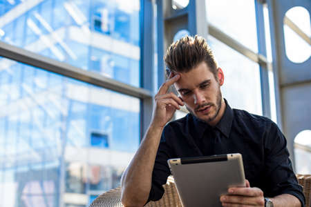 Thoughtful serious man online learning via touch pad, sitting in office interior. Male confident...