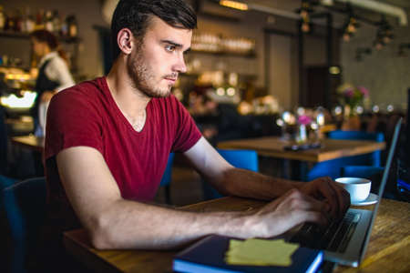 Handsome man skilled freelance copywriter keyboarding on laptop computer, sitting in modern coffee shop. Male professional content writer typing text on notebook during distance work in restaurantの写真素材