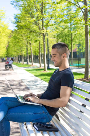 Joyful man university student preparing course work on laptop computer while sitting on a campus. Happy teenager working on netbook while resting in park in sunny summer day. Male using applicationsの写真素材