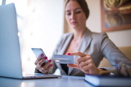 Business woman making online booking via mobile phone using debit credit card for payment, sitting with laptop computer in coffee shop. Female making online shopping store via cellphone gadgetの写真素材