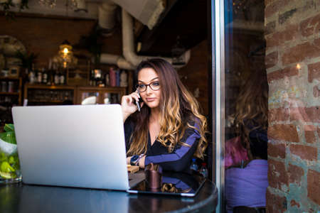 Woman freelance content manager taking via smartphone during distance work on laptop computer while sitting in restaurant during free time. Female having cellphone conversation during rest in cafeの写真素材