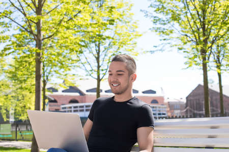 Smiling handsome man talking via online video call on netbook while sitting in park in sunny summer day. Cheerful male having distance work on laptop computer during rest outdoorsの写真素材