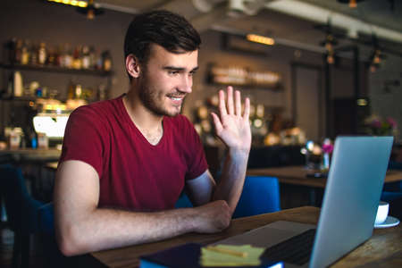 Happy smiling man waving hand hello during webcam conference via laptop computer, sitting in coffee shop interior. Cheerful male freelancer talking via video call on netbook, resting in restaurantの写真素材