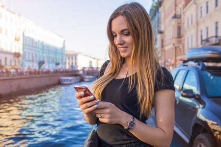 Young gorgeous woman online ordering via mobile phone while standing outdoors near Neva river background with copy space for promotional content. Pretty female traveler booking via cellphoneの写真素材