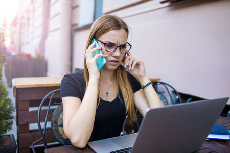 Serious angry woman professional business planning expert solves serious any questions with client via mobile phone and reading information in e-mail on laptop computer, sitting in restaurant outdoorsの写真素材