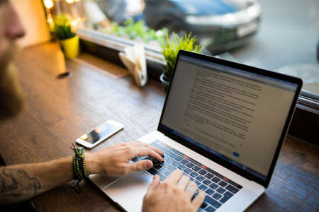 Close up hipster person having online training course via modern laptop computer while sitting in coffee shop. Cropped image man keyboarding on net-book preparing course work. Male writer using appsの写真素材