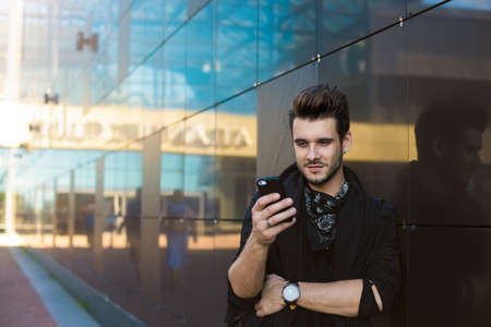 Young hipster guy dressed in casual stylish clothes watching video on mobile phone while standing...