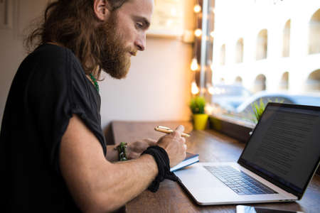 Serious bearded Sweden man with long hair having online training course via laptop computer, writing...