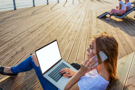 Female professional publication specialist having cellphone conversation during distance work on portable laptop computer with empty mockup copy space screen. Woman freelancer phoning via smartphoneの写真素材