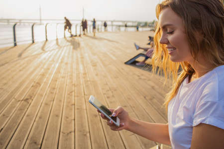 Happy smiling hipster girl using messenger on mobile phone during leisure time in the fresh air.Female reading text message on cellphone, sitting on wooden quay with copy space for promotional contentの写真素材