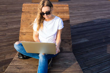 Caucasian female in stylish sunglasses having online education via laptop computer while sitting outdoors in sunny summer day. Hipster girl creative web content writer using notebook gadgetの写真素材