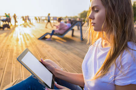 Hipster girl reading article in social network via touch pad while relaxing on wooden embankment in...