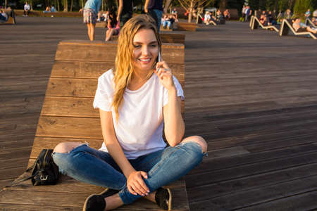 Young happy hipster girl talking via cell telephone while sitting outdoors on bench in park in sunny...