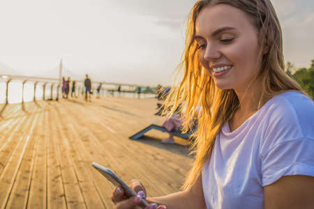 Happy smiling hipster girl watching video in social network via smartphone while sitting outdoors near copy space for promotional content. Cheerful female sending sms via cellphone during walking timeの写真素材
