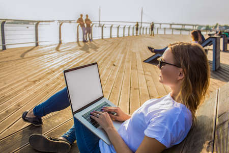 Happy smiling fashionable hipster girl in stylish sunglasses keyboarding on portable laptop computer with empty mockup copy space screen background for promotional content. Female chatting via netbookの写真素材
