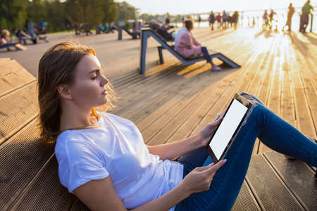 Attractive woman reading news in internet via modern touch pad with empty mock up copy space screen background for promotional content while sitting on bench in park in sunny summer evening.の写真素材