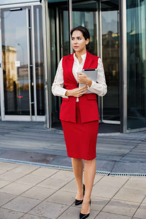Young successful business woman dressed in luxury formal wear holding touch pad while standing outside company while waiting partner. Female boss standing against enterpriseの写真素材