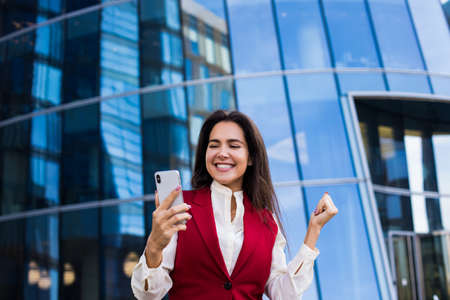 Successful happy smiling woman entrepreneur reading on mobile phone lucky good news while standing outside contemporary business skyscraper building.の写真素材