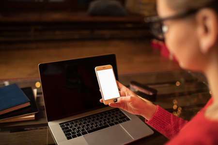 View from behind of a hipster girl holding modern mobile phone with empty mockup copy space screen background for your advertising text message while sitting with laptop computer at the table in homeの写真素材