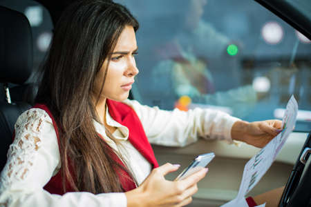 Frustrated business woman reading paper documents with financial graphs with bad result and using mobile phone for checking information while sitting in luxury automobile. Upset female CEO in carの写真素材