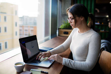 Serious woman in stylish casual wear and spectacles professional business content writer searching information in internet via pc laptop computer, sitting in coffee shop. Hipster girl using appsの写真素材