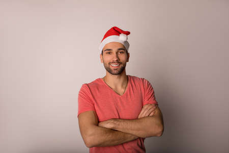 Portrait of a confident cheerful man in red Canta Clause hat standing with crossed arms isolated in studio against background with copy space for promotional content and smiling for cameraの写真素材