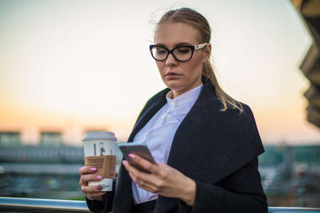 Serious woman broker checking e-mail on mobile phone while standing with take away coffee outdoors in evening time. Female college student reading sms on cell telephone.の写真素材