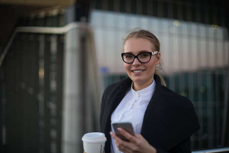 Portrait of a cheerful woman entrepreneur in stylish spectacles, white shirt and autumn coat holding in hands mobile phone and cup of coffee and smiling for camera while standing outside companyの写真素材