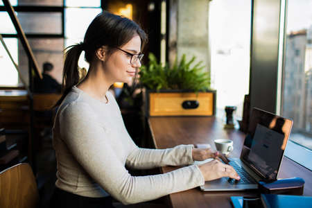 Woman smart publication specialist creating web site via modern laptop computer, sitting in coffee shop. Female skilled copywriter distance working on netbook during recreation time in co-working cafeの写真素材