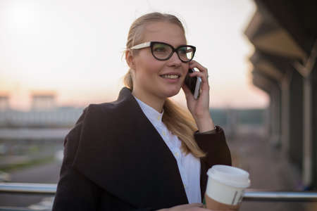 Happy smiling woman office worker in stylish spectacles and casual coat having pleasant mobile phone conversation while standing with take away coffee outdoors in evening time during work breakの写真素材
