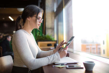 Serious woman advertiser in fashionable glasses searching article on web site via touch pad while...