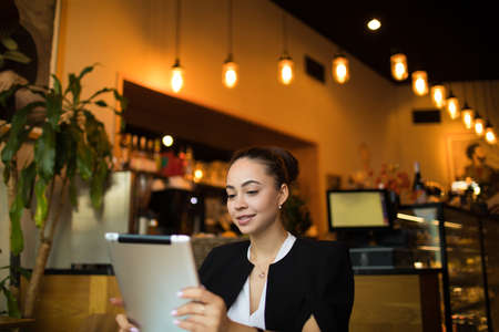 Smiling woman professional lawyer reading article on touch pad while sitting in restaurant before meeting with client. Female university student online banking via digital tablet computerの写真素材