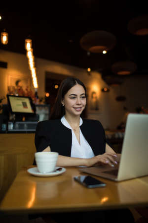 Young beautiful smiling woman successful publication specialist typing article on laptop computer while sitting in coffee shop during recreation time. Happy female copywriter having training courseの写真素材