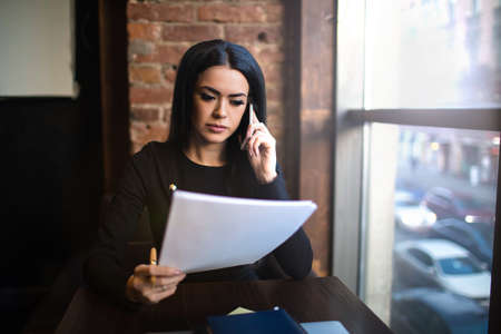 Serious woman financier exploring paper documents with new contract, sitting in restaurant after work day. Female skilled business owner reading summary during cellphone conversationの写真素材