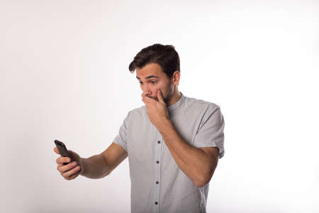 Shocked man covers his mouth with his hand while reading bad news in sms on cell telephone while standing isolated in studioの写真素材