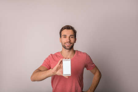 Handsome cheerful hipster guy in casual t-shirt holding mobile phone with empty mock up copy space...
