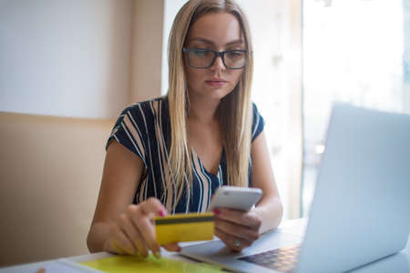 Young woman manager in spectacles using debit credit card for online payment via mobile phone during...