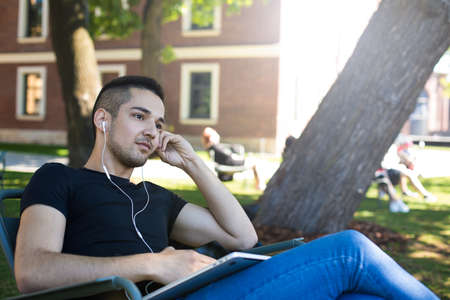 Thoughtful man dreaming and listening to music via headphones while sitting in park in summer day during recreation time in weekend. Pensive hipster boy listening audio training via earphonesの写真素材