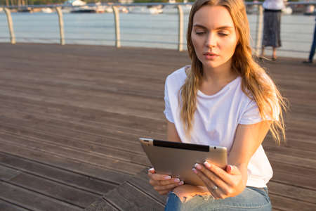 Gorgeous woman online booking via portable touch pad gadget while relaxing outdoors during summer vacations. Thoughtful college female reading e-book via digital tablet, resting on quay in sunny dayの写真素材