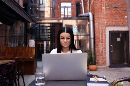 Woman in trendy spectacles clever college student having online learning via laptop computer, sitting near modern building outdoors. Female intelligent journalist accounting via netbook gadgetの写真素材