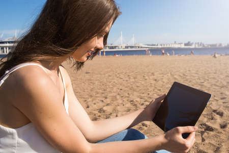 Smiling female publication specialist reading fashion news via portable touch pad computer while relaxing on the beach in sunny day during summer vacation. Woman online booking via digital gadgetの写真素材